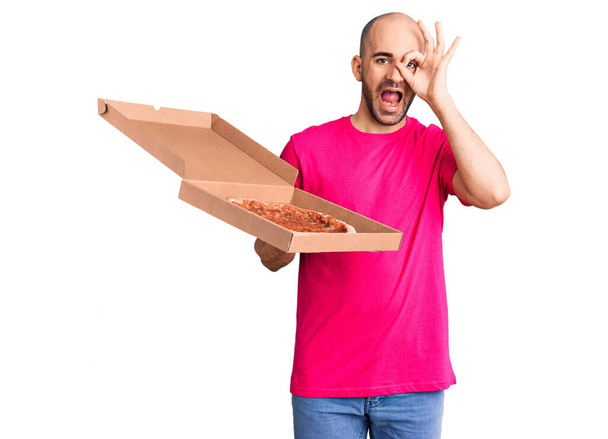 Fiatal jóképű férfi kezében szállítás pizza kartondoboz mosolyogva boldog csinál rendben jel kézzel a szemen keresztül néz az ujjak  - Fotó, kép