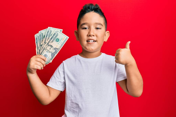 Pequeño niño hispano sosteniendo billetes de 20 dólares sonriendo feliz y positivo, pulgar hacia arriba haciendo excelente y signo de aprobación  - Foto, imagen