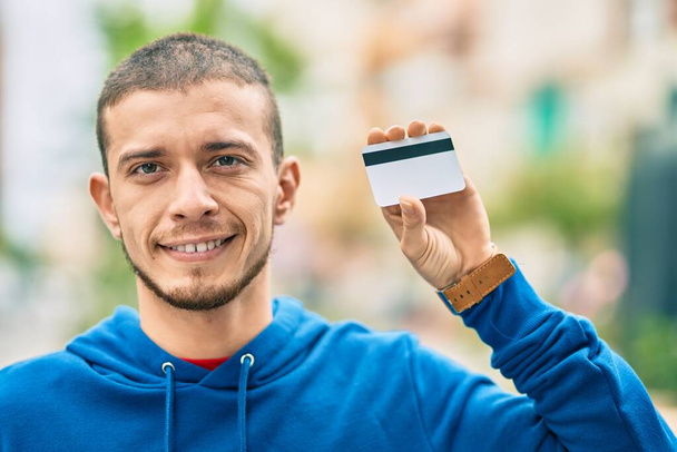 Giovane uomo ispanico sorridente felice in possesso di carta di credito in città - Foto, immagini