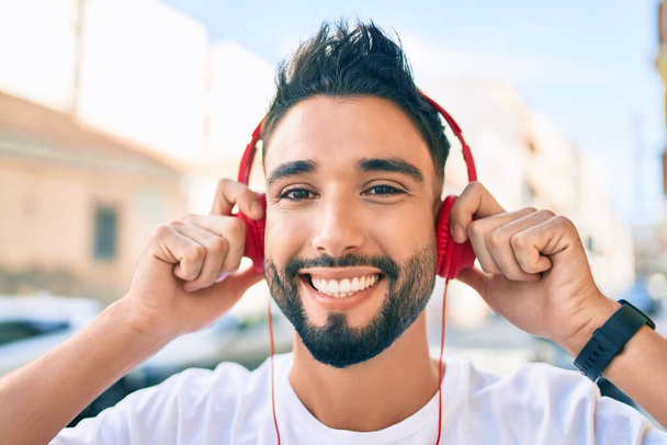 Νεαρός Άραβας χαμογελά χαρούμενος χρησιμοποιώντας ακουστικά με τα πόδια στην πόλη. - Φωτογραφία, εικόνα