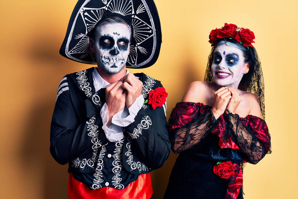 Jeune couple portant le jour mexicain du costume mort sur jaune rire nerveux et excité avec les mains sur le menton regardant vers le côté  - Photo, image