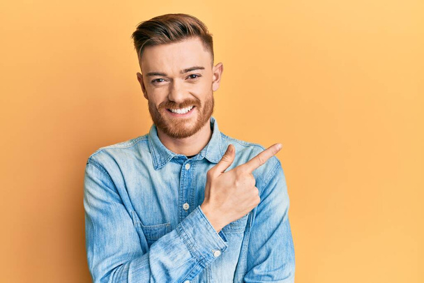 Junger rothaariger Mann in lässigem Jeanshemd lächelt fröhlich und zeigt mit Hand und Finger zur Seite  - Foto, Bild