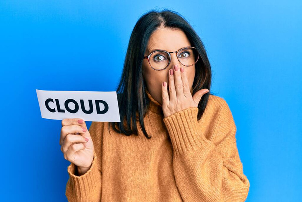 Middle age brunette woman holding paper with cloud word covering mouth with hand, shocked and afraid for mistake. surprised expression  - Фото, изображение