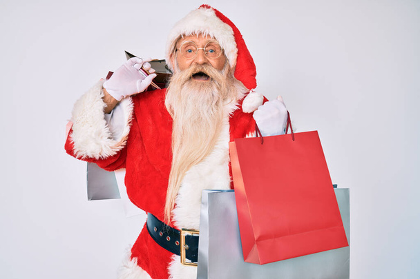 Old senior man with grey hair and long beard wearing santa claus costume holding shopping bags smiling and laughing hard out loud because funny crazy joke.  - Фото, изображение