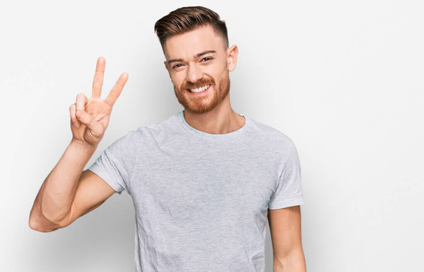Young redhead man wearing casual grey t shirt showing and pointing up with fingers number two while smiling confident and happy.  - Photo, Image