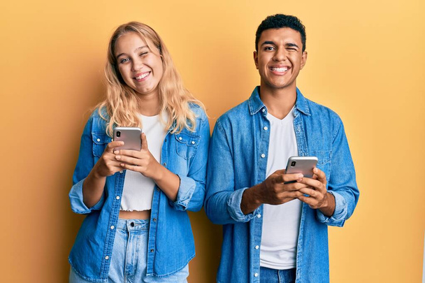 Jeune couple interracial en utilisant smartphone clin d'oeil en regardant la caméra avec une expression sexy, gai et heureux visage.  - Photo, image