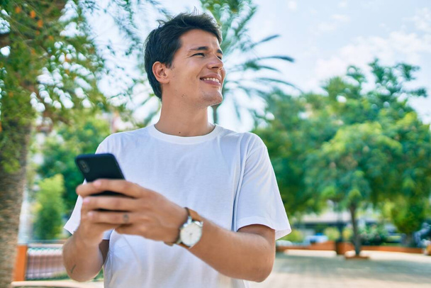 Mladý kavkazský muž usměvavý šťastný pomocí smartphonu ve městě. - Fotografie, Obrázek