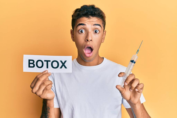 Young handsome african american man holding botox banner and needle afraid and shocked with surprise and amazed expression, fear and excited face.  - Photo, Image