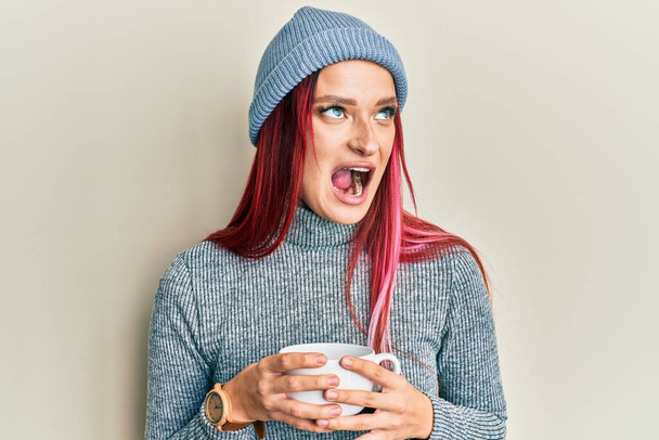 Kafkasyalı genç bir kadın kış şapkası takıyor ve bir fincan sıcak kahve içiyor kızgın ve öfkeli bir şekilde bağırıyor, öfkeyle bağırıyor. öfke ve agresif konsept.  - Fotoğraf, Görsel