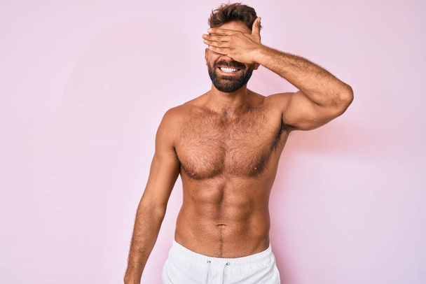 Young hispanic man standing shirtless smiling and laughing with hand on face covering eyes for surprise. blind concept.  - 写真・画像