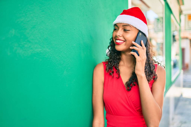 Young african american girl wearing christmas hat talking on the smartphone leaning on the wall. - Foto, Imagen