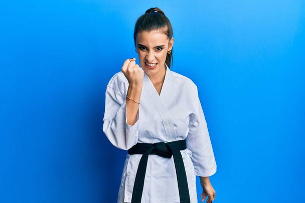 Beautiful brunette young woman wearing karate fighter uniform with black belt angry and mad raising fist frustrated and furious while shouting with anger. rage and aggressive concept.  - 写真・画像