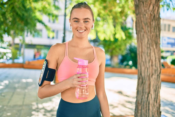 Young blonde sportswoman doing exercise drinking bottle of water at the park - 写真・画像