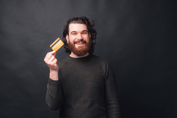 A happy young bearded man is holding a credit card looking at the camera near a black wall - Fotografie, Obrázek