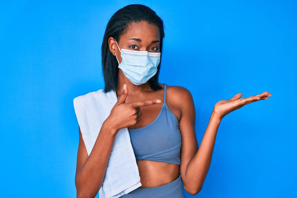 Молода афроамериканка, одягнена в спортивне лаяння і медична маска, вражена і посміхається перед камерою, представляючи руками і вказуючи пальцем..  - Фото, зображення