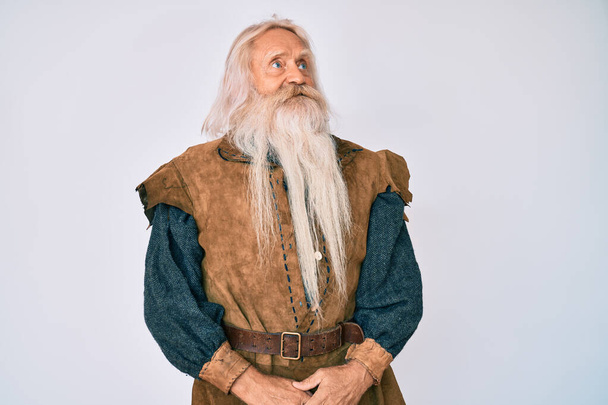 Vieil homme âgé aux cheveux gris et à la barbe longue portant un costume traditionnel viking détournant les yeux avec le sourire sur le visage, expression naturelle. riant confiant.  - Photo, image