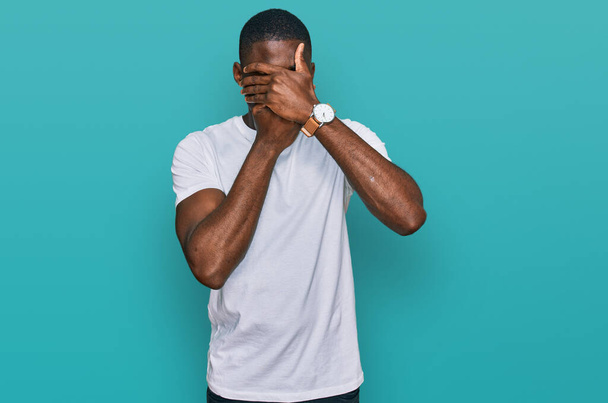 Joven hombre afroamericano vistiendo casual camiseta blanca que cubre los ojos y la boca con las manos, sorprendido y sorprendido. ocultar emociones  - Foto, imagen