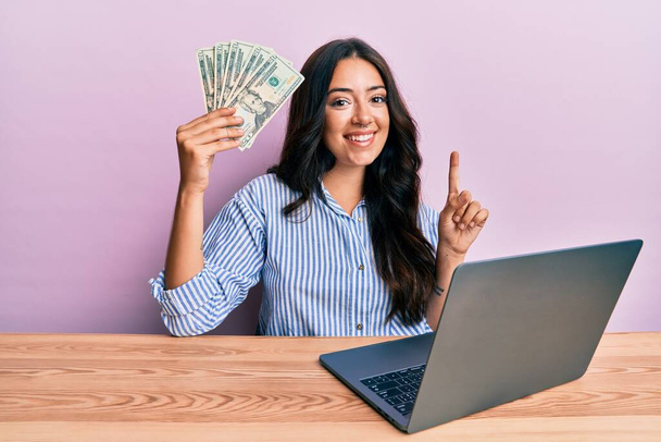 Όμορφη μελαχρινή νεαρή γυναίκα που εργάζεται στο γραφείο κρατώντας δολάρια χαμογελώντας με μια ιδέα ή ερώτηση δείχνοντας το δάχτυλο με χαρούμενο πρόσωπο, νούμερο ένα  - Φωτογραφία, εικόνα