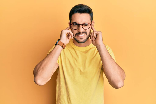 Junger hispanischer Mann in lässiger Kleidung und Brille, der die Ohren mit den Fingern mit genervtem Gesichtsausdruck vor lauter Musik verdeckt. Gehörloses Konzept.  - Foto, Bild