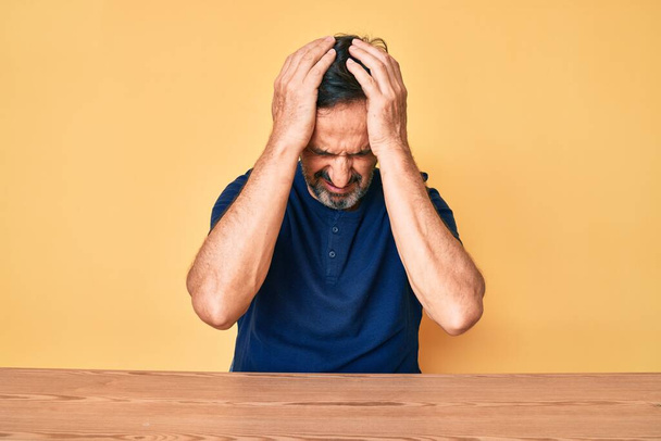 Hombre hispano de mediana edad con ropa casual sentado en la mesa sufriendo de dolor de cabeza desesperado y estresado porque el dolor y la migraña. manos en la cabeza.  - Foto, Imagen