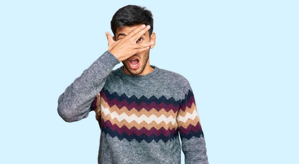 Jonge knappe man draagt casual winter trui gluren in shock bedekking gezicht en ogen met de hand, kijken door vingers met beschaamde uitdrukking.  - Foto, afbeelding