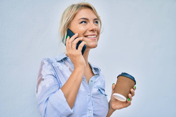 Young blonde woman smiling happy talking on the smartphone and drinking take away coffee at the city. - Foto, Imagen