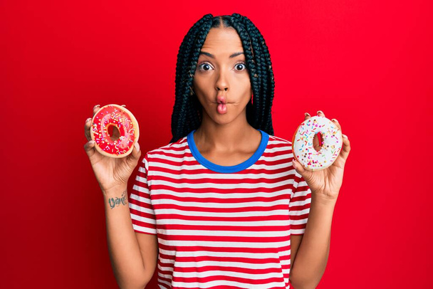 Beautiful hispanic woman holding tasty colorful doughnuts making fish face with mouth and squinting eyes, crazy and comical.  - Foto, afbeelding