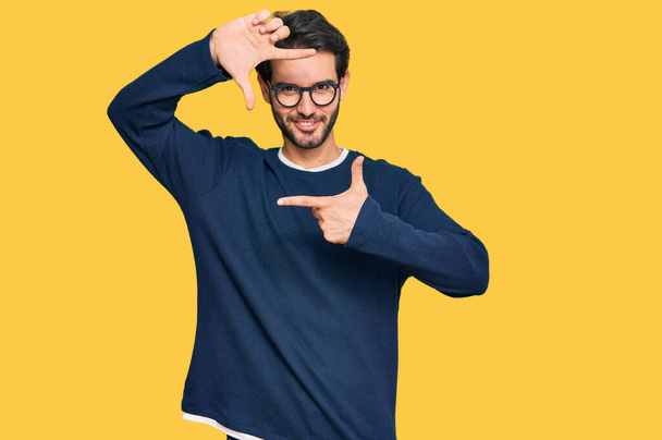Joven hombre hispano vistiendo ropa casual y gafas sonrientes haciendo montura con manos y dedos con cara feliz. concepto de creatividad y fotografía.  - Foto, Imagen