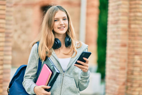 Όμορφη καυκάσιος φοιτητής έφηβος χαμογελά χαρούμενος χρησιμοποιώντας smartphone στην πόλη. - Φωτογραφία, εικόνα