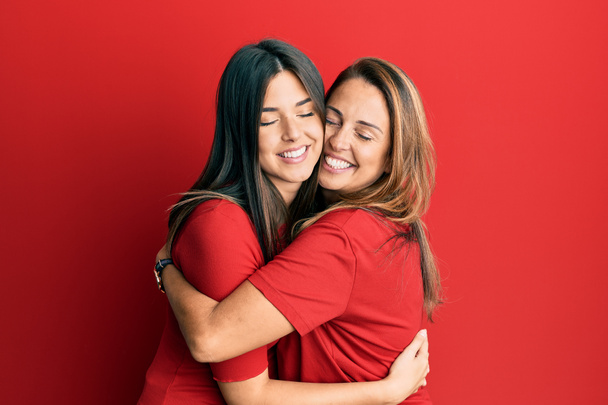 Die hispanische Familie aus Mutter und Tochter trägt lässige Kleidung vor rotem Hintergrund und umarmt sich glücklich und positiv, lächelt selbstbewusst. Selbstliebe und Selbstfürsorge  - Foto, Bild