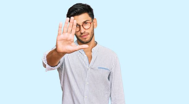 Jonge knappe man in casual kleding en bril doet stoppen met zingen met de palm van de hand. waarschuwingsuitdrukking met negatief en ernstig gebaar op het gezicht.  - Foto, afbeelding