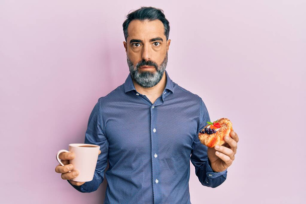 Мужчина средних лет с бородой и седыми волосами, пьющий кофе и поедающий выпечку в шокирующем лице, скептически и саркастично выглядящий, удивленный открытым ртом  - Фото, изображение