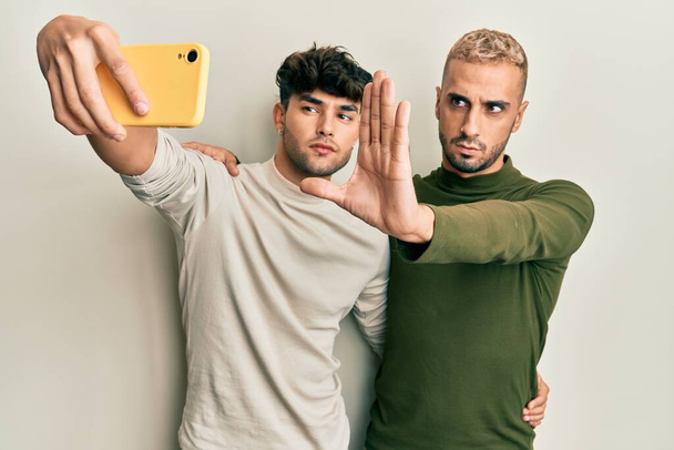 Homoseksueel gay paar staan samen het nemen van een selfie foto met smartphone met open hand doen stop teken met ernstige en zelfverzekerde expressie, verdediging gebaar  - Foto, afbeelding