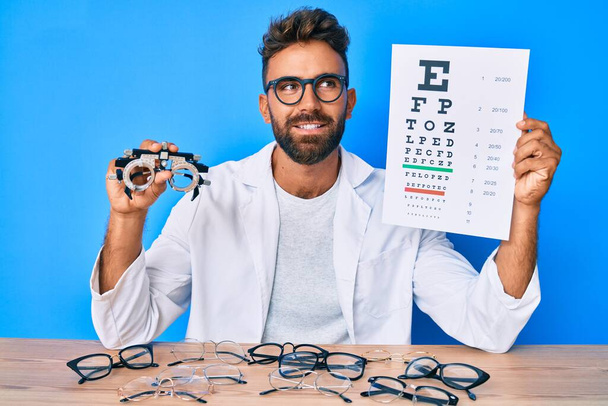 Νεαρός Ισπανός με γυαλιά οπτομετρίας και χαρτί με γράμματα που χαμογελούν κοιτώντας στο πλάι και κοιτώντας αλλού σκεπτόμενος.  - Φωτογραφία, εικόνα