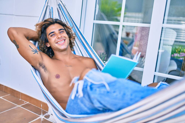 Giovane uomo ispanico rilassato libro di lettura sdraiato sull'amaca in terrazza. - Foto, immagini