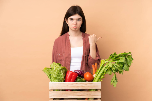 Fiatal farmer lány frissen szedett zöldségekkel egy dobozban boldogtalan és oldalra mutató - Fotó, kép