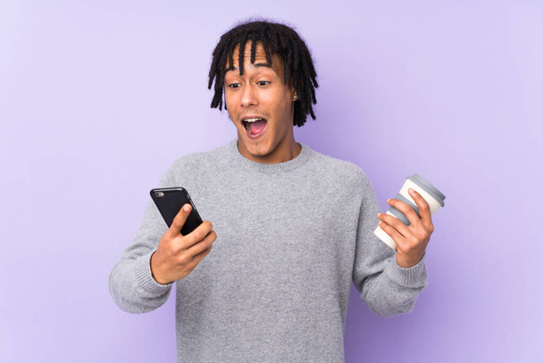 Jeune homme afro-américain isolé sur fond violet tenant du café à emporter et un mobile - Photo, image