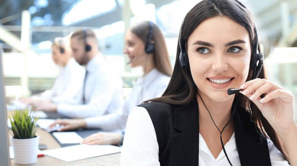 Female customer support operator with headset and smiling, with collegues at background. - Fotoğraf, Görsel
