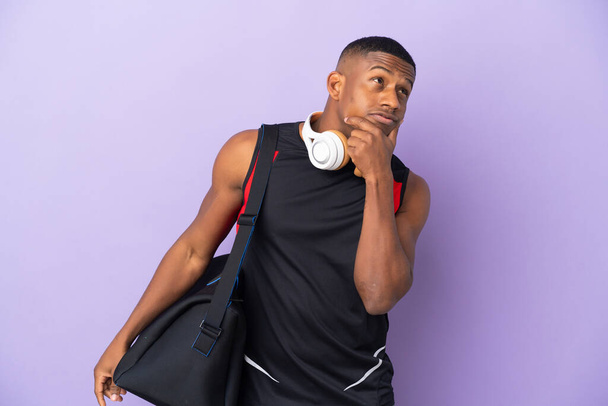 Mladý sportovní latinský muž se sportovní taškou izolované na fialovém pozadí s pochybnostmi - Fotografie, Obrázek