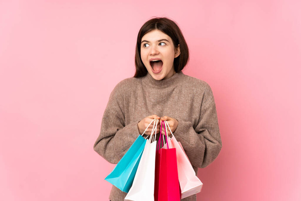 Young Ukrainian teenager girl over isolated pink background holding shopping bags and surprised - Фото, изображение