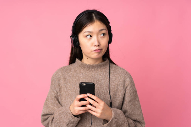 Young asian woman isolated on pink background listening music with a mobile and thinking - Valokuva, kuva