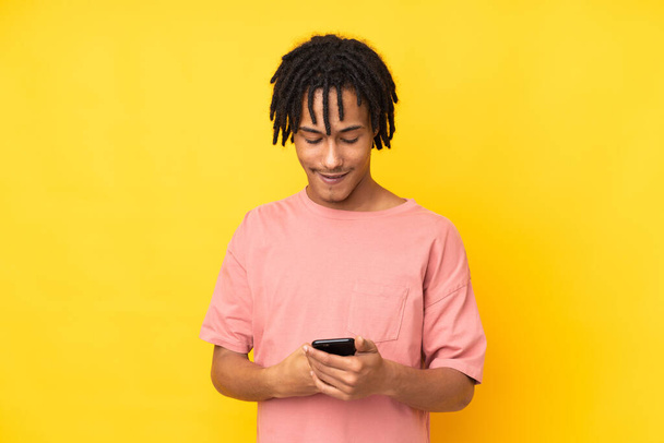 Молодой африканский американец, изолированный на желтом фоне, отправляет сообщение с мобильным телефоном - Фото, изображение