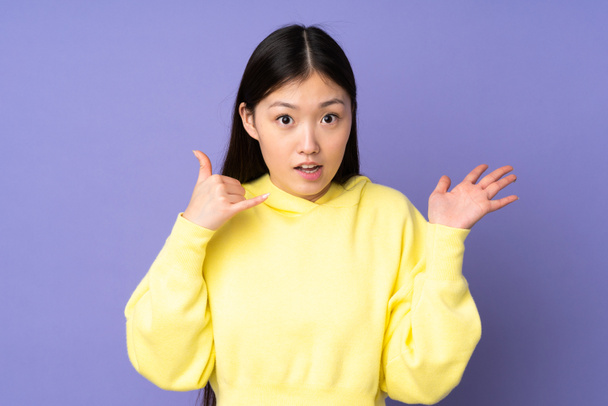 Giovane donna asiatica isolata su sfondo viola fare gesto del telefono e dubitare - Foto, immagini