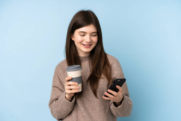 Junge ukrainische Teenager Mädchen trägt einen Pullover über isoliertem blauem Hintergrund mit Kaffee zum Mitnehmen und ein Handy - Foto, Bild