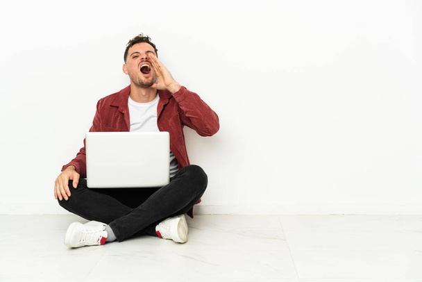 młody przystojny biały mężczyzna siedzieć w w na podłodze z laptop krzycząc z usta szeroko otwarte - Zdjęcie, obraz