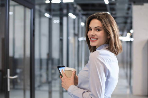 Businesswoman walking along the office corridor with paper cup and smartphone - Photo, Image