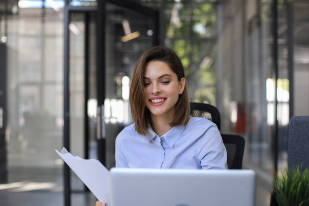 Attractive cheerful business woman checking paper documents in office, working on laptop - Фото, изображение