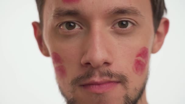 Yüzünde kırmızı ruj izi olan yakışıklı bir Avrupalı adamın portresi oral seks jestini yakından gösteriyor. Erkek yasak seks sembolü yapar, gülümser, beyaz arka planda sürpriz yapar. Sadakatsizlik kavramı - Video, Çekim