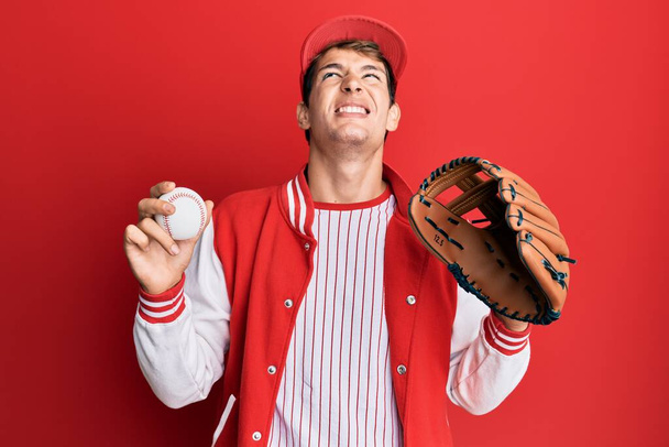 Handsome caucasian man wearing baseball uniform holding golve and ball angry and mad screaming frustrated and furious, shouting with anger looking up.  - Fotó, kép
