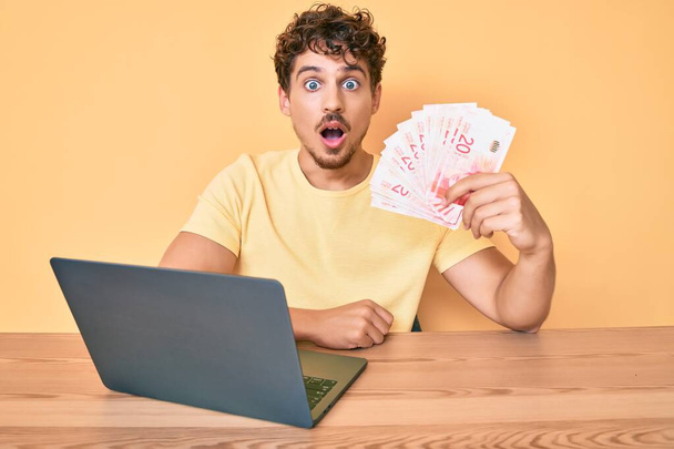 młody biały mężczyzna z kręconymi włosami siedzi na stole pracując z laptopem i trzymając israel sykle banknoty przestraszony i zdumiony z otwartymi ustami dla zaskoczenia, niedowierzanie twarz  - Zdjęcie, obraz